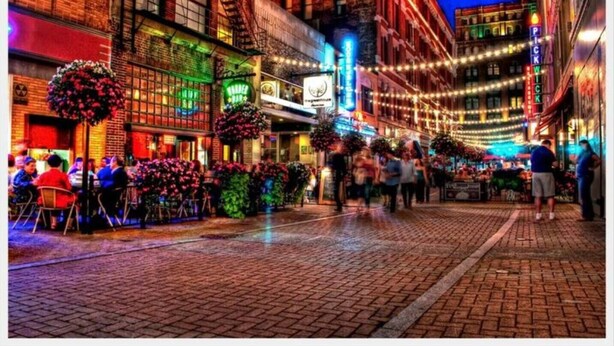 Una calle animada con edificios de ladrillo, mesas al aire libre y luces colgantes.