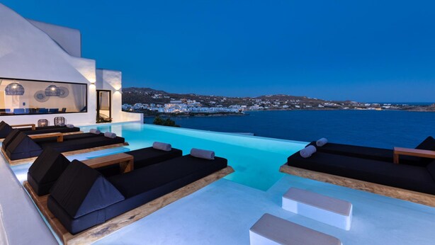Una piscina infinita moderna con sillones de descanso y vistas a un pueblo costero al anochecer.