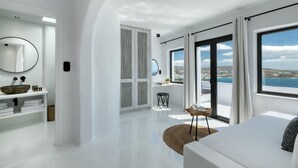 Un dormitorio moderno con una cama grande, vistas al mar y un baño con lavamanos y espejo blancos.