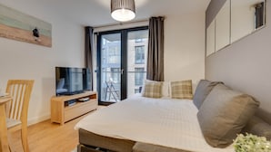 Ein modernes Schlafzimmer mit einem großen Bett, einem Fernseher auf einem Holzgestell, einem Balkon mit Blick auf Gebäude und einem Bild an der Wand.