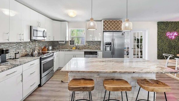 Una cucina moderna con mobili bianchi, una grande isola con sgabelli, elettrodomestici in acciaio inossidabile e illuminazione a sospensione.