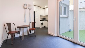 Een compacte keuken met een wasmachine, een eethoek met twee stoelen en een kleine tafel, en een schuifdeur naar een balkon met uitzicht op een gazon.