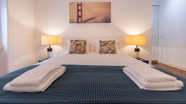 Een hotelkamer met een groot bed, twee bedlampen, decoratieve kussens, een schilderij van een brug en een blauw tapijt met structuur.