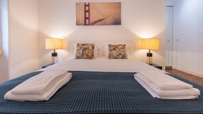 Habitación de hotel con una cama grande, dos lámparas de noche, almohadones decorativos, un cuadro de un puente y una alfombra azul con textura.