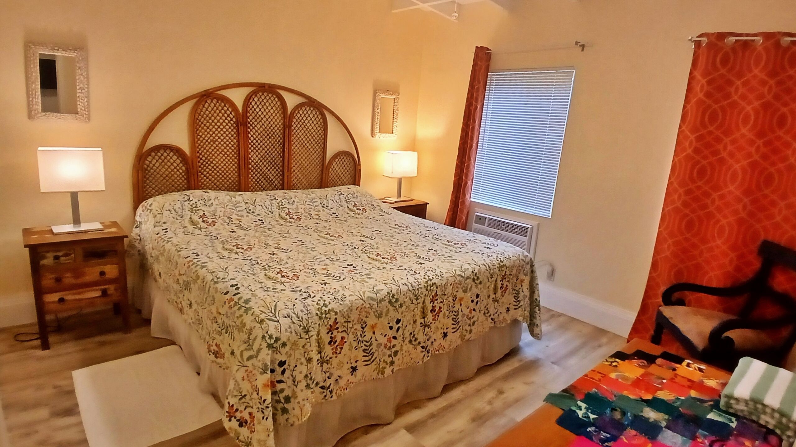 A bedroom with a bed, two bedside tables, a window with blinds, a door with a curtain, and a small mirror on the wall.