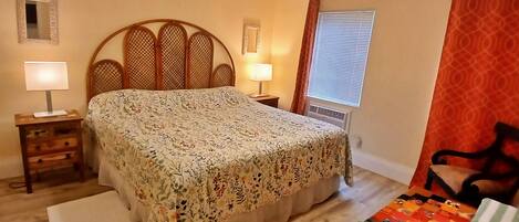 A bedroom with a bed, two bedside tables, a window with blinds, a door with a curtain, and a small mirror on the wall.