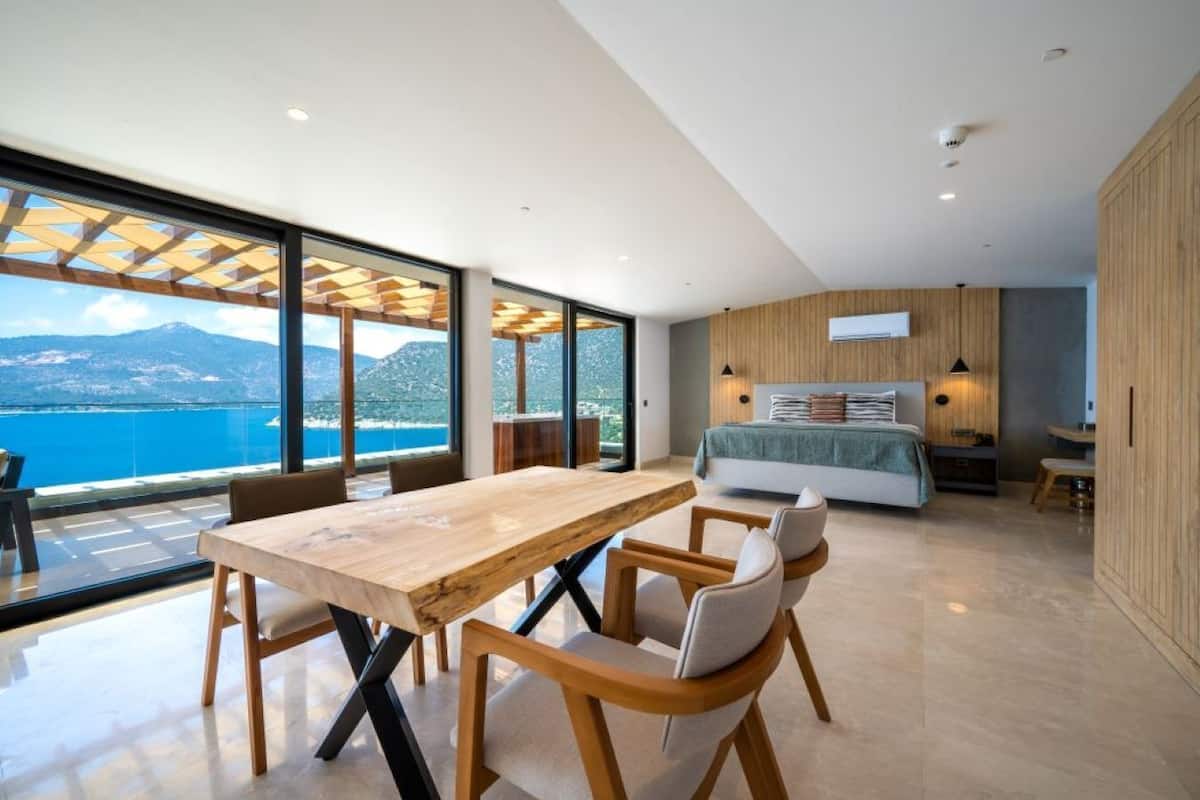 Una habitación de hotel moderna con una amplia mesa de comedor de madera, asientos cómodos, una cama con colcha verde y vistas al mar.
