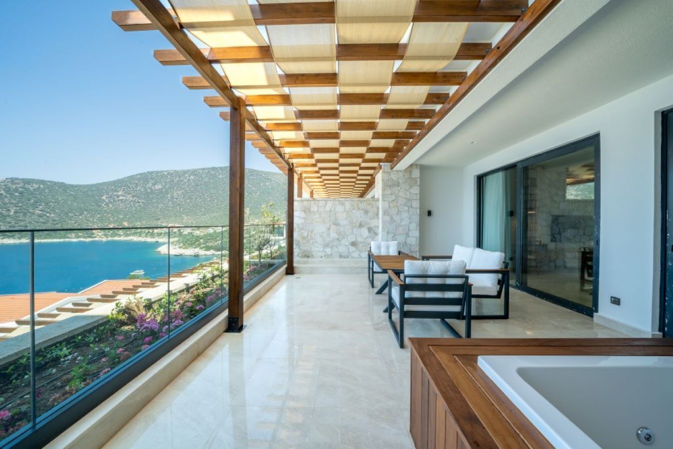 Un balcón con vista al mar, una pérgola de madera, una zona de estar con cojines blancos y una bañera grande.