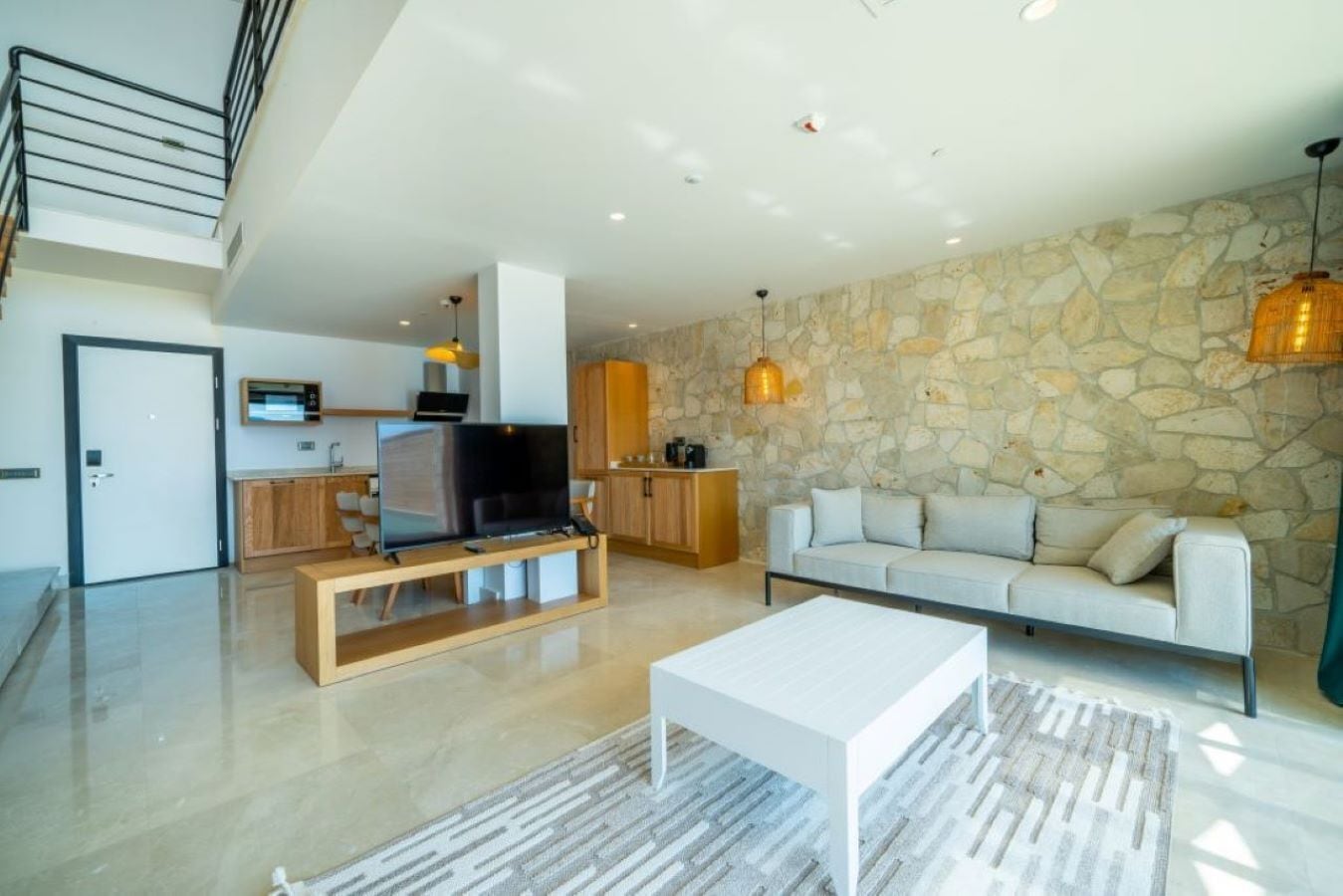 A modern living room with a flat-screen TV, a sofa, and a coffee table. There is a kitchen area in the background with wooden cabinets and a stone wall.