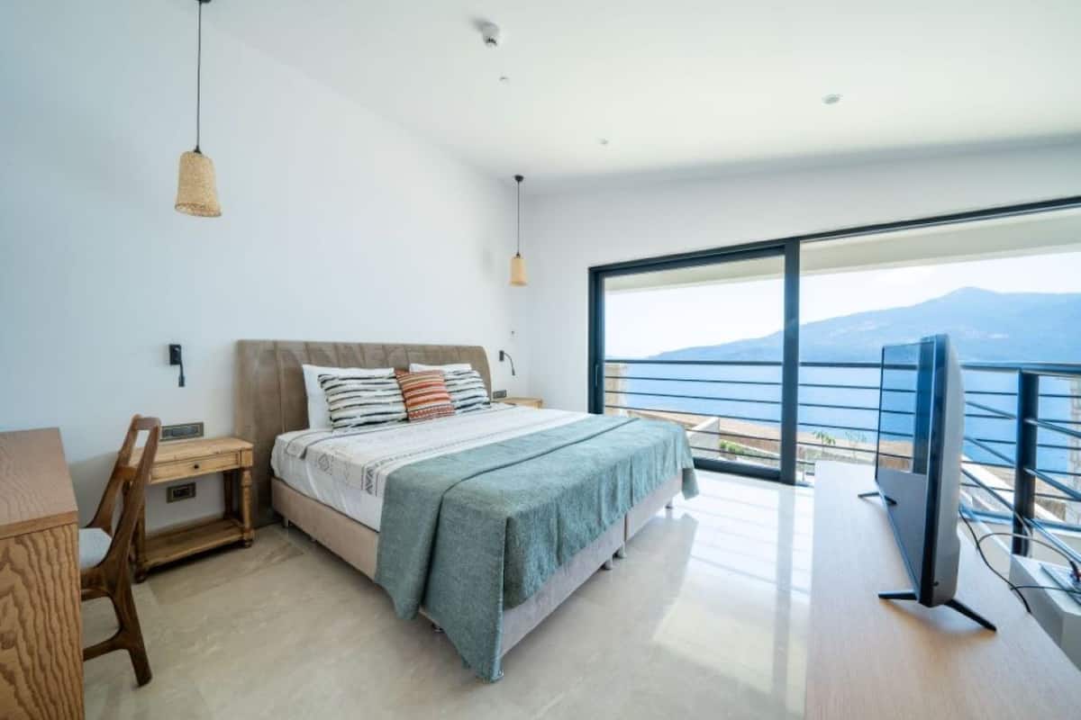 Un dormitorio moderno con una cama grande, un escritorio y un televisor. Tiene un balcón con vistas al mar y a las montañas.