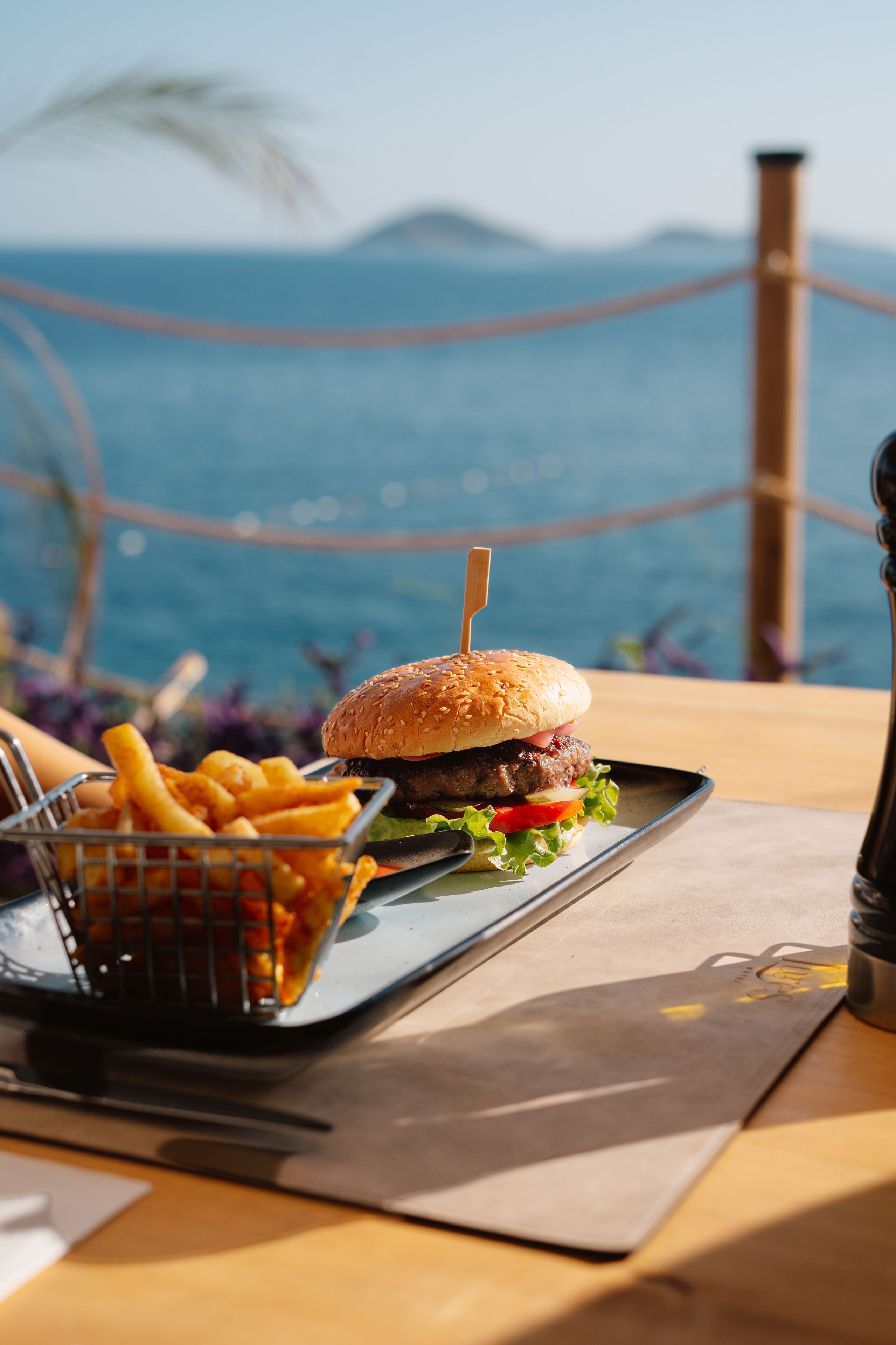 Un hamburger accompagné de frites, servi sur un plateau, avec une vue sur la mer et les montagnes en arrière-plan.