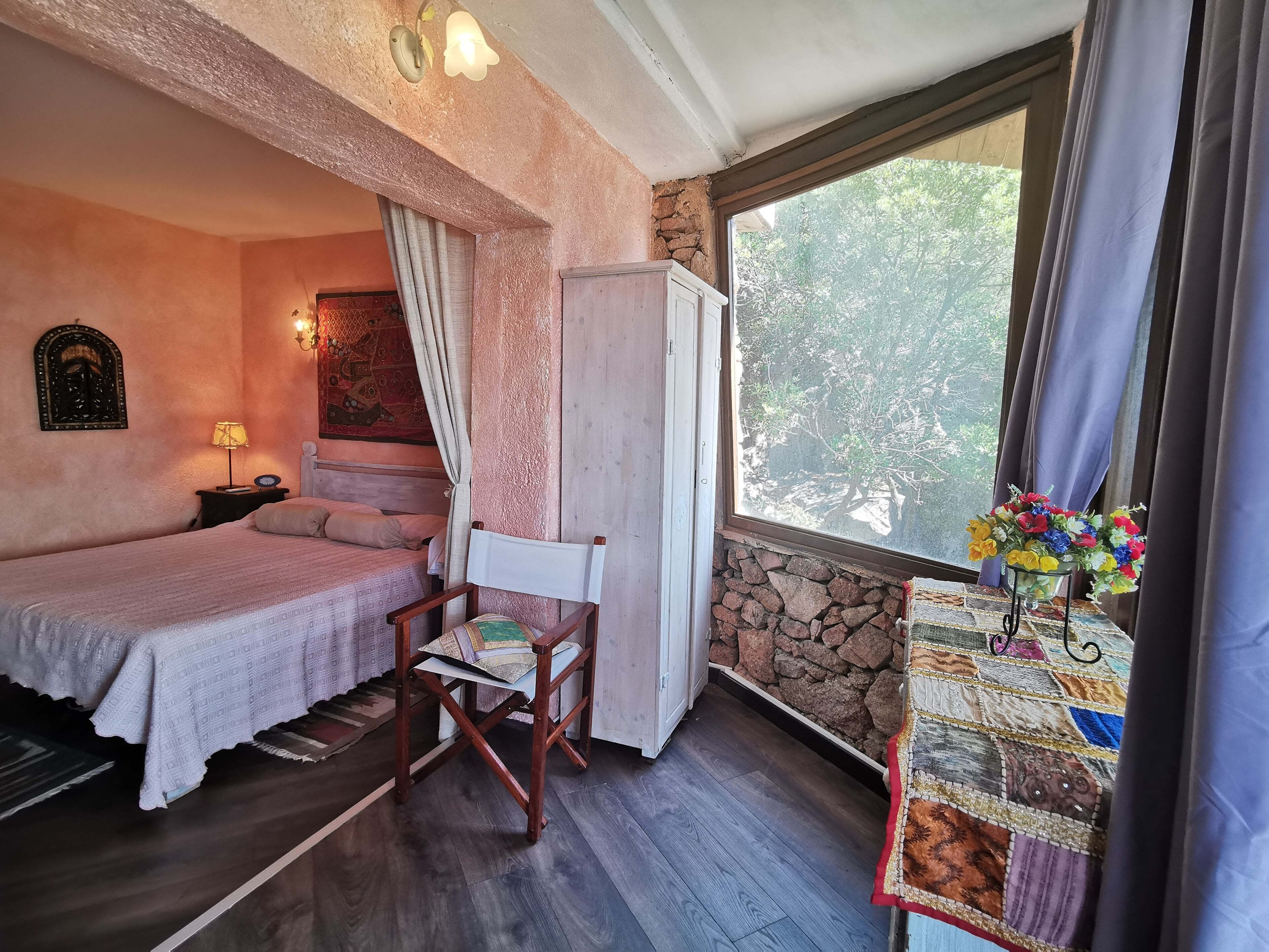Un dormitorio con una cama, una silla, una mesita con colcha, una ventana con cortinas y una pared de piedra.
