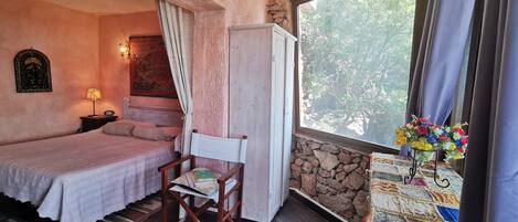 Un dormitorio con una cama, una silla, una mesita con colcha, una ventana con cortinas y una pared de piedra.