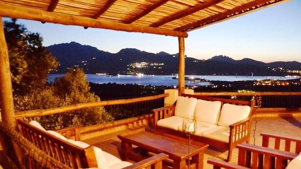 A covered outdoor seating area with wooden furniture and a view of a mountainous landscape and a body of water at dusk. - Villa Vista, from the terrace a stunning view to  Costa Smeralda and the Islands (Sardegna)