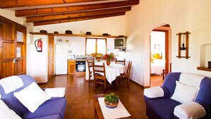 A cozy living room with wooden beams, a dining area with a table and chairs, and a kitchen in the background.