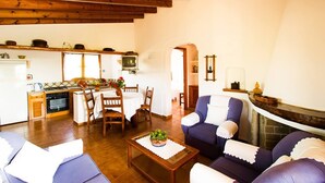 A cozy living room with a fireplace, wooden beams, a dining area with a table and chairs, and a kitchen with a stove and cabinets.