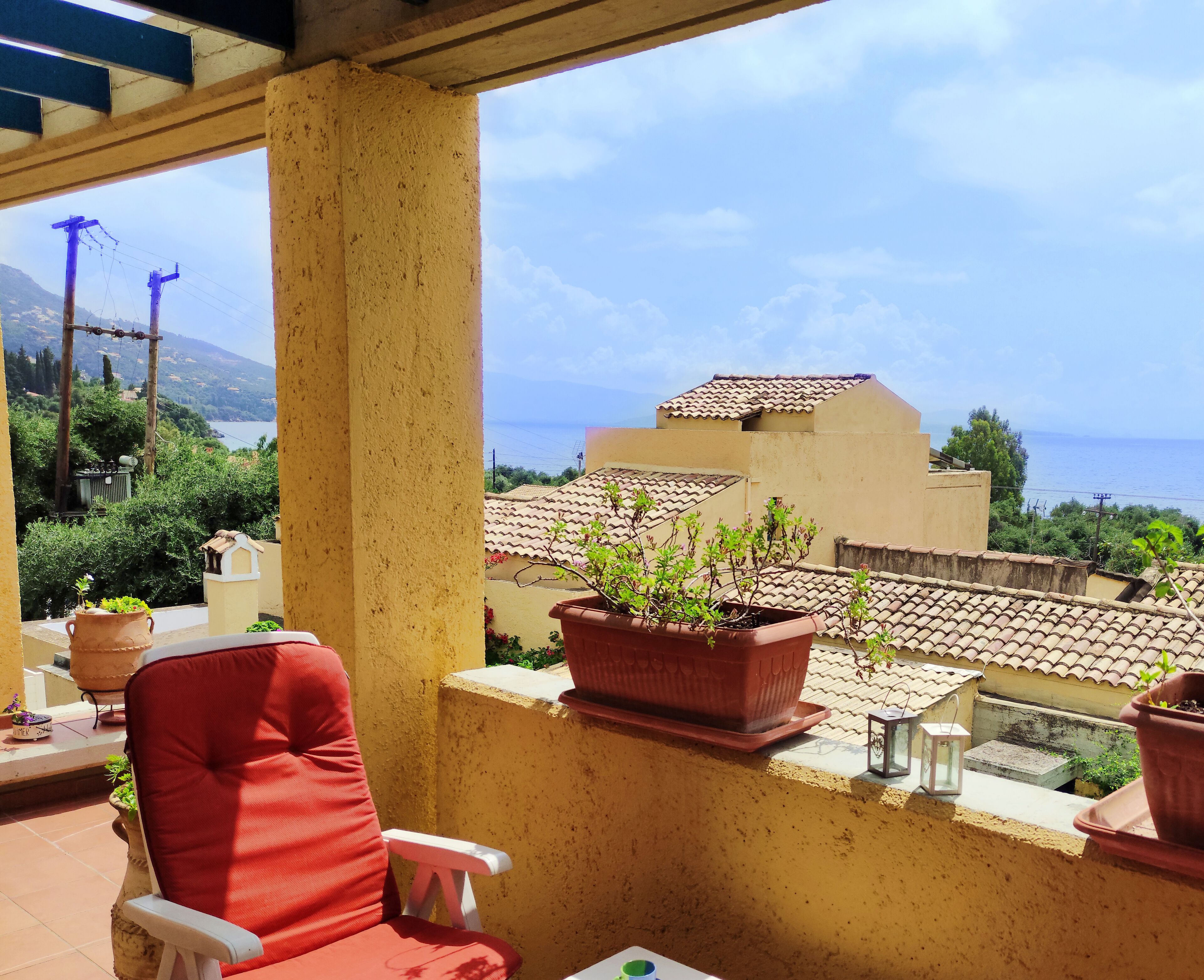 A balcony with a red chair, terracotta pots with plants, and a view of a coastal area with buildings and mountains in the background.