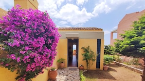 A yellow building with a tiled roof, a door, and a small garden area with potted plants and a flowering bush. - Mira El Mar by Corfuescapes (Corfu)
