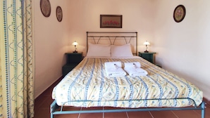 A neatly made bed with a floral-patterned bedspread, two pillows, and two folded towels on a metal frame. There are two bedside tables with lamps, and a framed floral artwork above the bed. - Mira El Mar by Corfuescapes (Corfu)