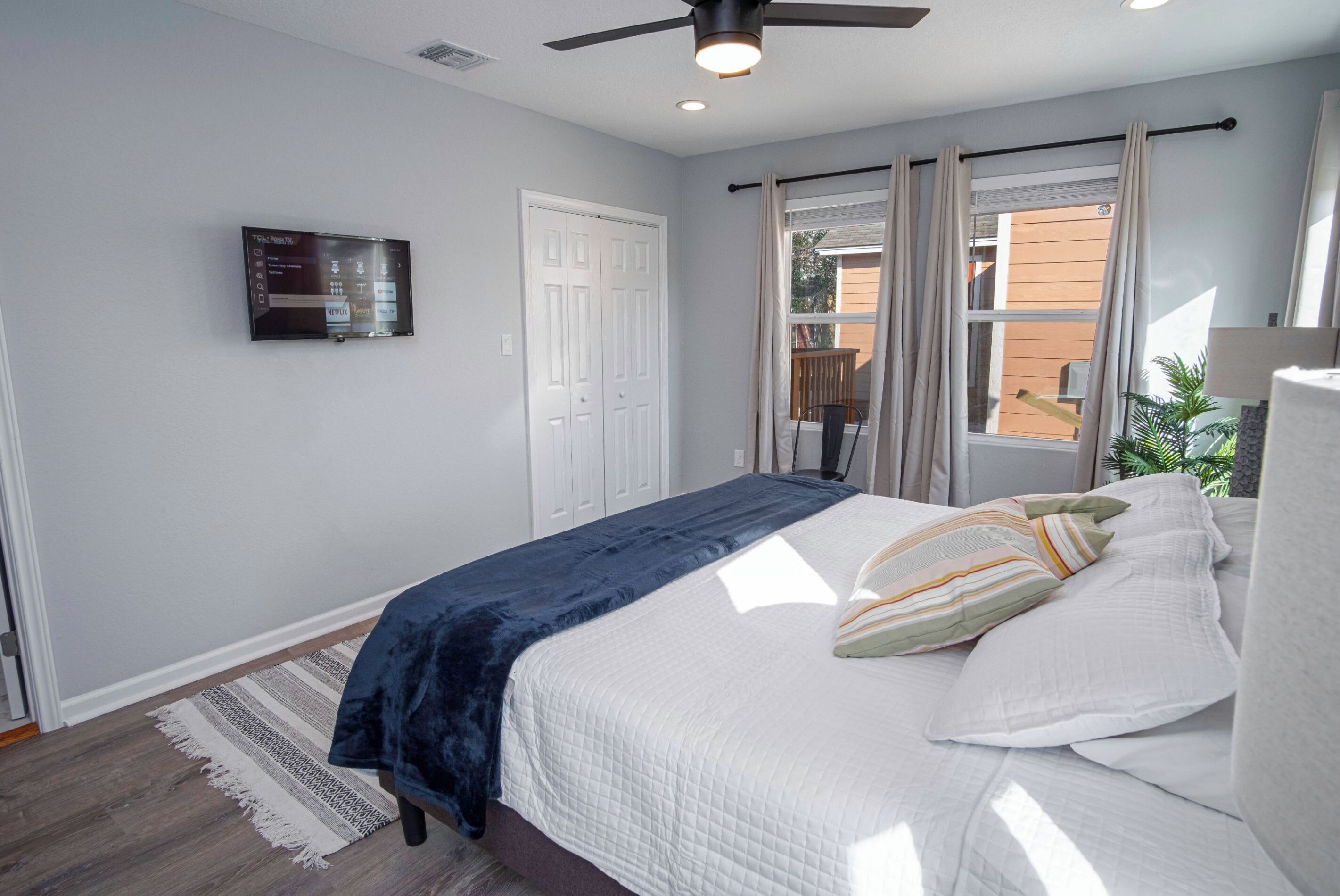 A bedroom with a bed, a ceiling fan, a TV mounted on the wall, and a window with curtains.