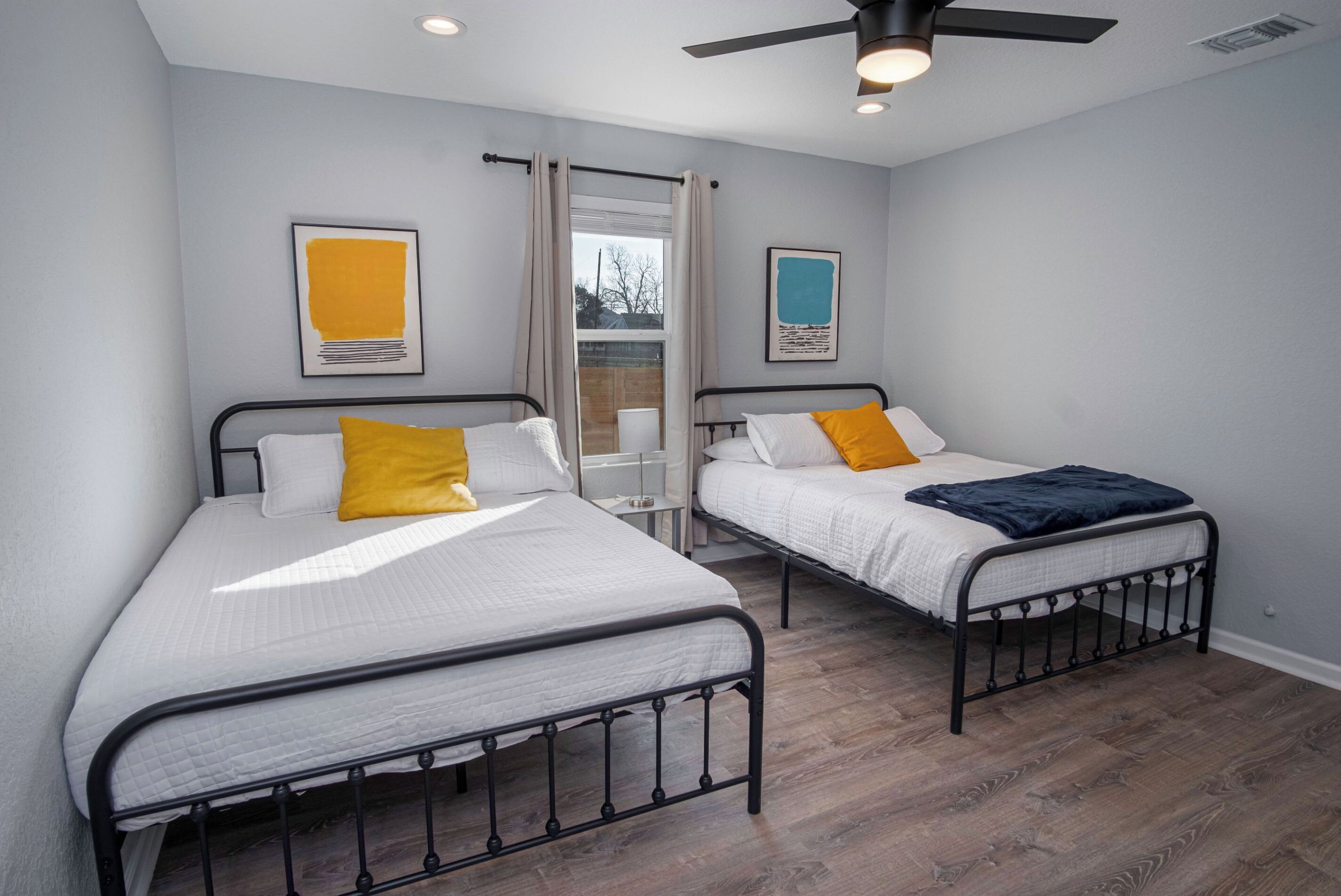 A room with two beds, each with a metal frame and white bedding. There are two framed artworks on the walls, a ceiling fan, and a window with curtains.