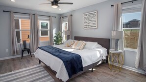 Une chambre à coucher avec un lit, un ventilateur de plafond, une fenêtre avec des rideaux, une chaise et une petite table.