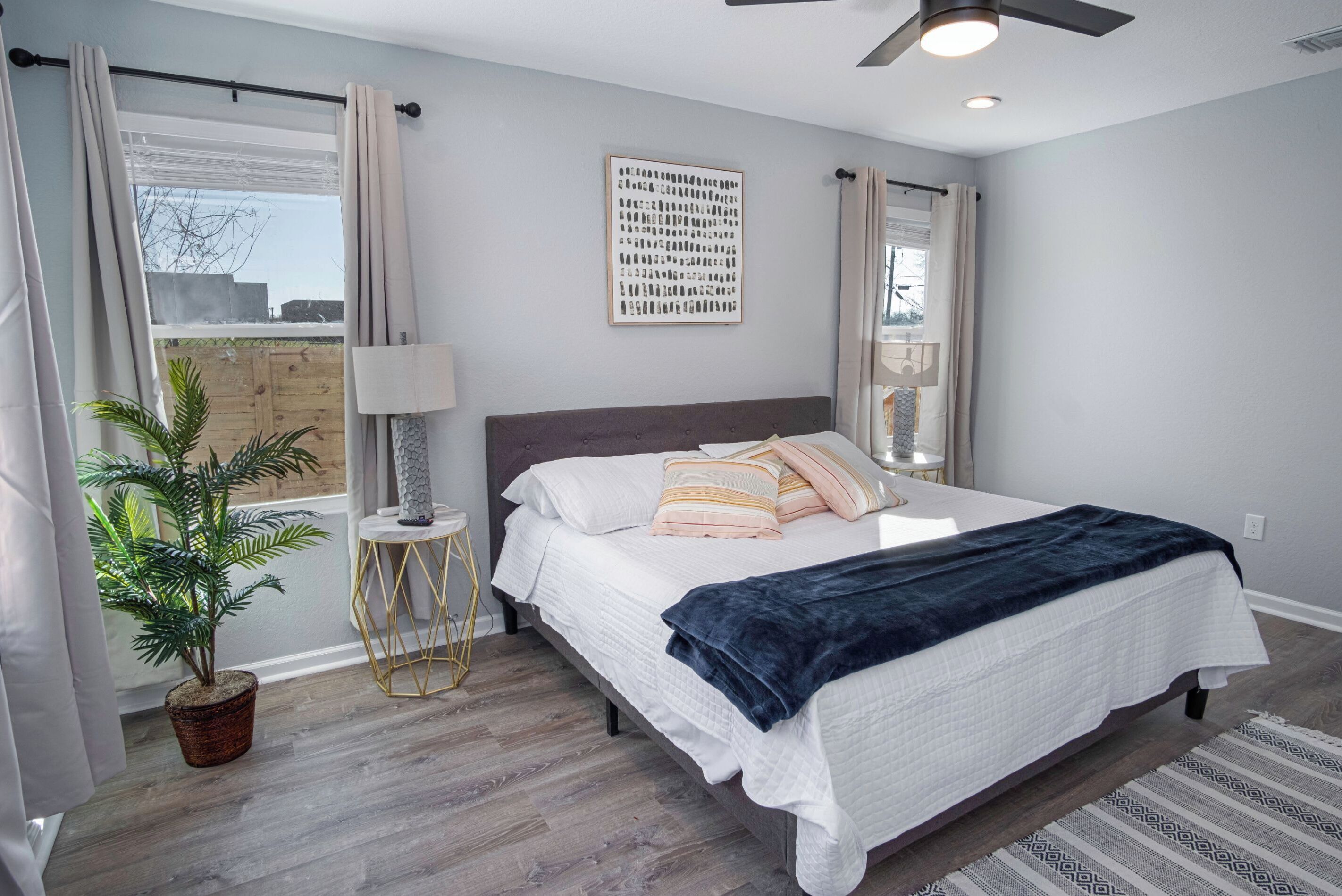 Une chambre à coucher avec un lit, un ventilateur de plafond, une lampe fixée au mur, une plante en pot et une fenêtre avec des rideaux.