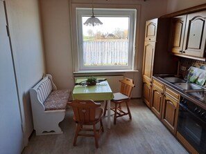 A kitchen with wooden cabinets, a dining table with chairs, a bench, and a window with a view of a fence and trees. - Relax in the iodine-rich North Sea air in the first floor apartment Deichblick (Friedrichskoog)