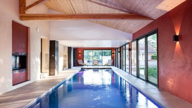 A modern indoor swimming pool with clear glass walls, wooden beams, and a red accent wall. - Le Cottage du Parc (Chambéon)