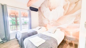 A hotel room with two beds, a large floral mural, a window with a view of greenery, and a small bedside table with a lamp. - Le Cottage du Parc (Chambéon)