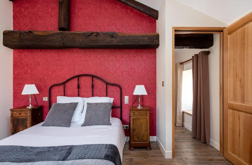 A bedroom with a red textured wall, a wooden ceiling beam, a bed with white bedding, two bedside tables with lamps, and a wooden door.