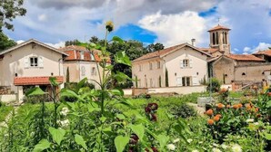 A row of traditional buildings with a central tower, surrounded by lush greenery and flowering plants. - Le Cottage du Parc (Chambéon)