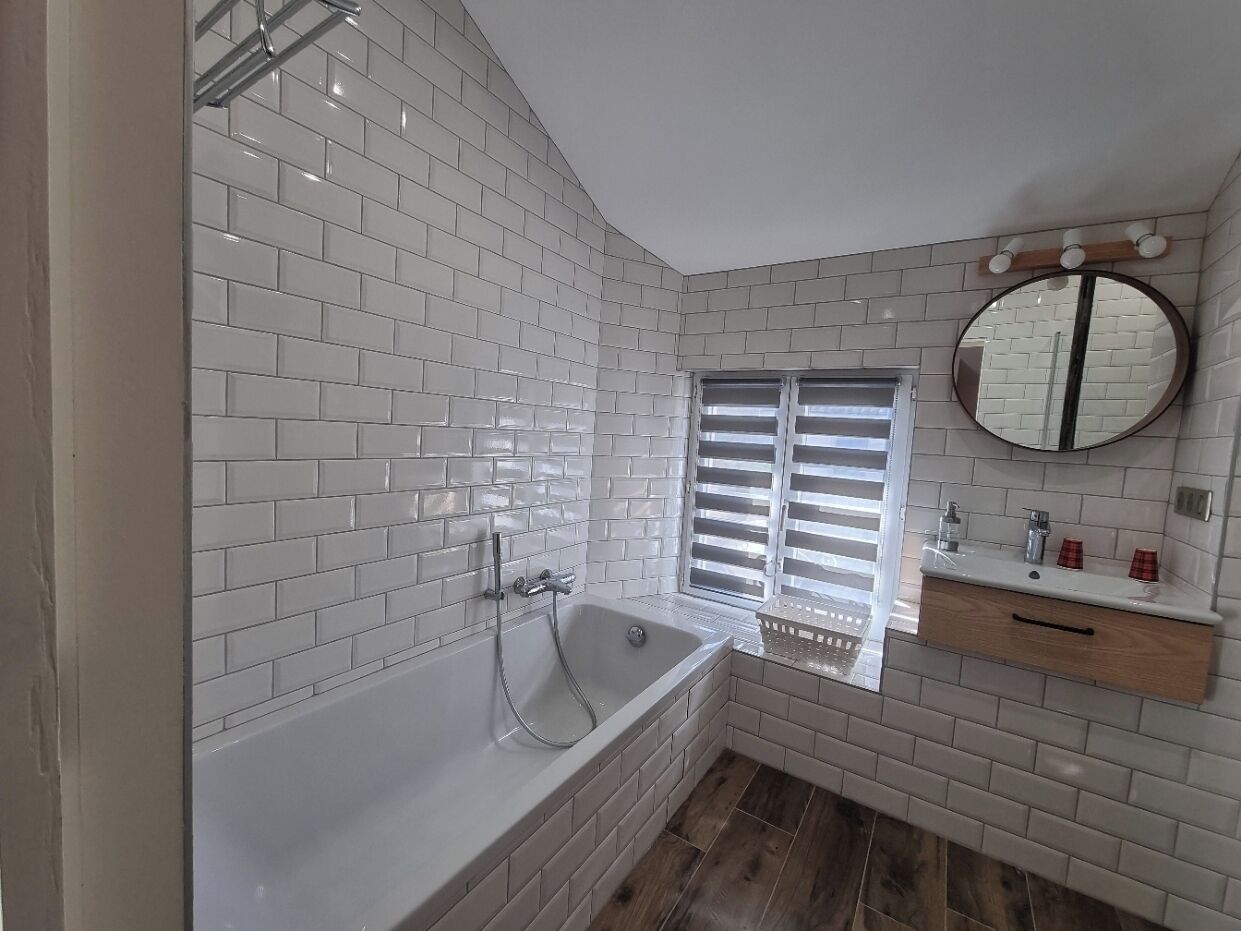 Un baño con ducha de azulejos blancos, tocador de madera con espejo y ventana con persianas.
