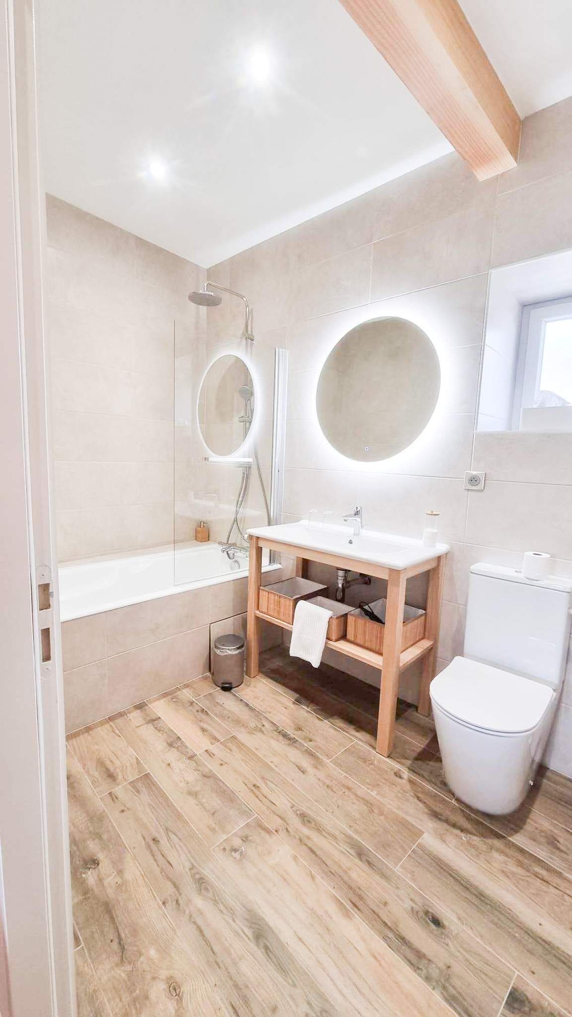 A modern bathroom with a wooden vanity, a bathtub, a toilet, and two round mirrors.