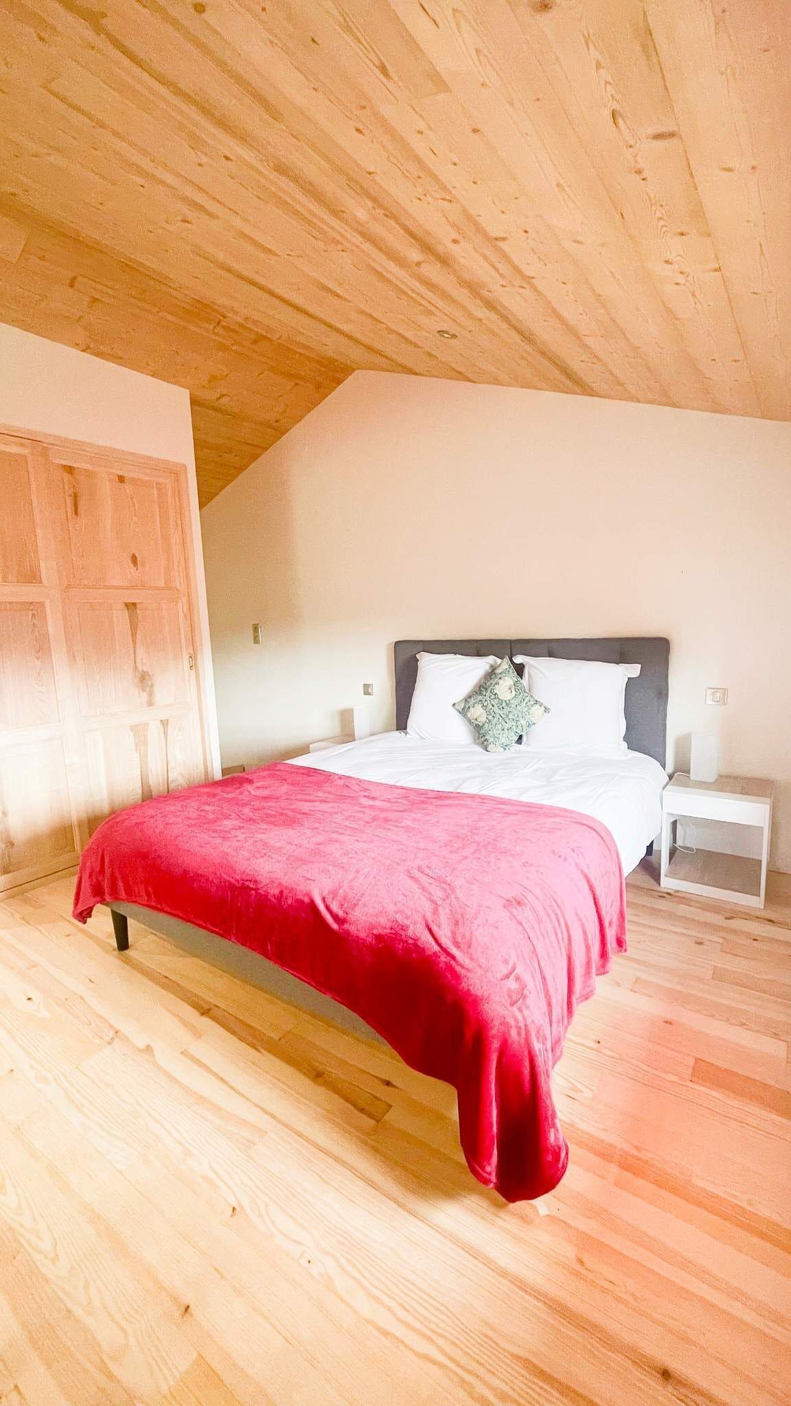 Quarto com teto de madeira, cama com manta vermelha, roupa de cama branca e mesa de cabeceira com abajur.
