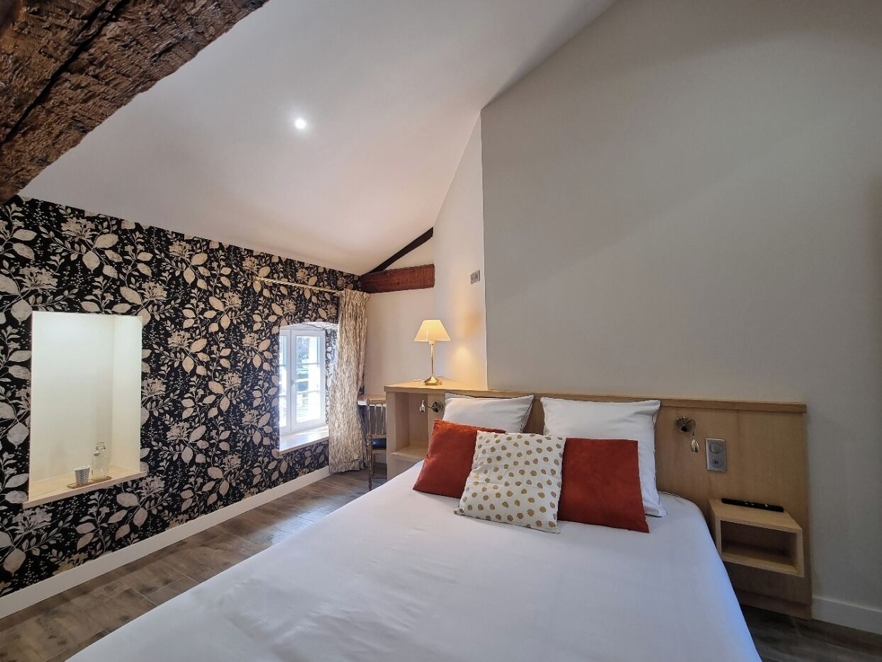 A bedroom with a large bed, a nightstand, a lamp, a window with curtains, and a wall with a floral pattern.