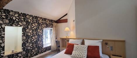 A bedroom with a large bed, a nightstand, a lamp, a window with curtains, and a wall with a floral pattern.