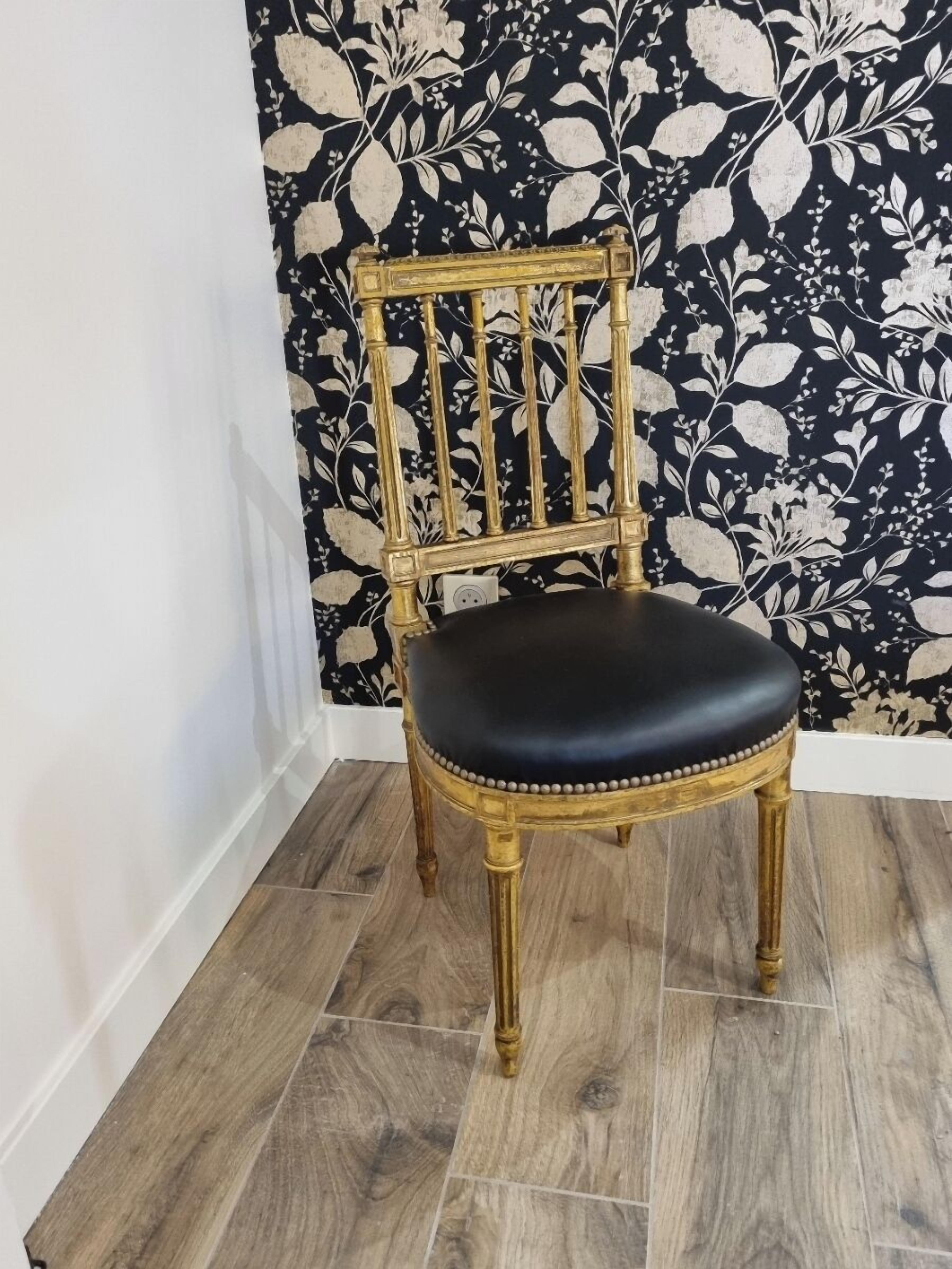 Una silla de madera con asiento negro y detalles dorados, sobre un piso de madera frente a una pared con estampado floral.