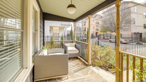 Een overdekt balkon in de open lucht met rieten meubilair, een hanglamp en uitzicht op de straat en de gebouwen.