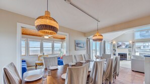 Un vaste espace dédié à la salle à manger, avec une grande table en bois et de nombreuses chaises, de grands luminaires suspendus en fibres tressées, et une vue sur l’océan grâce à de grandes fenêtres.