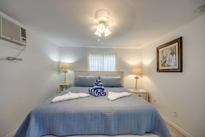 Une chambre à coucher comprenant un lit, deux lampes de chevet, un ventilateur de plafond, un appareil de climatisation, une fenêtre avec des rideaux et un tableau encadré accroché au mur.
