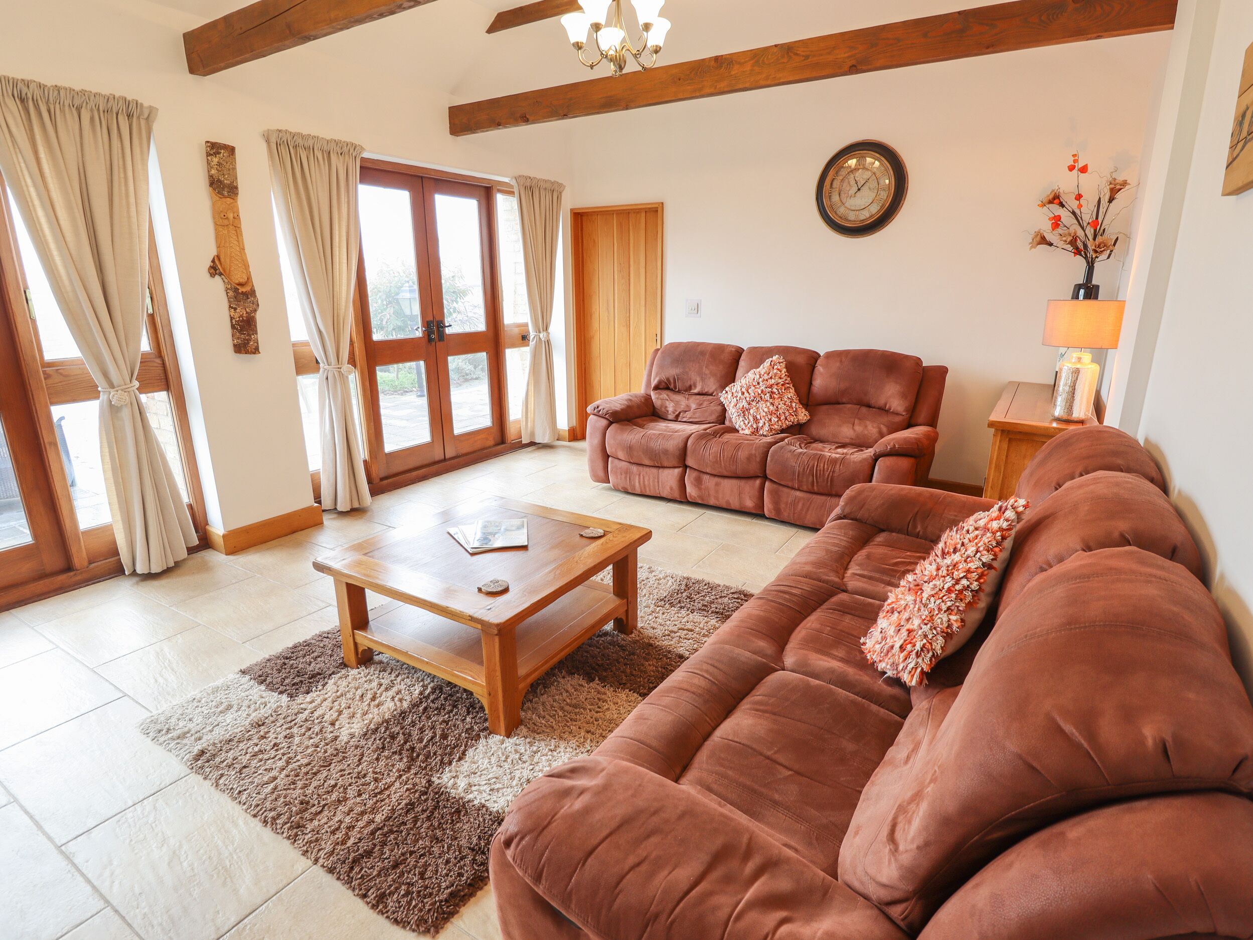 Ein Wohnzimmer mit braunem Sofa, einem Holztisch und einem Teppich. Außerdem eine Holztür und ein Fenster mit Vorhängen.