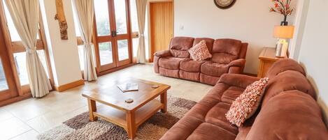 Ein Wohnzimmer mit braunem Sofa, einem Holztisch und einem Teppich. Außerdem eine Holztür und ein Fenster mit Vorhängen.