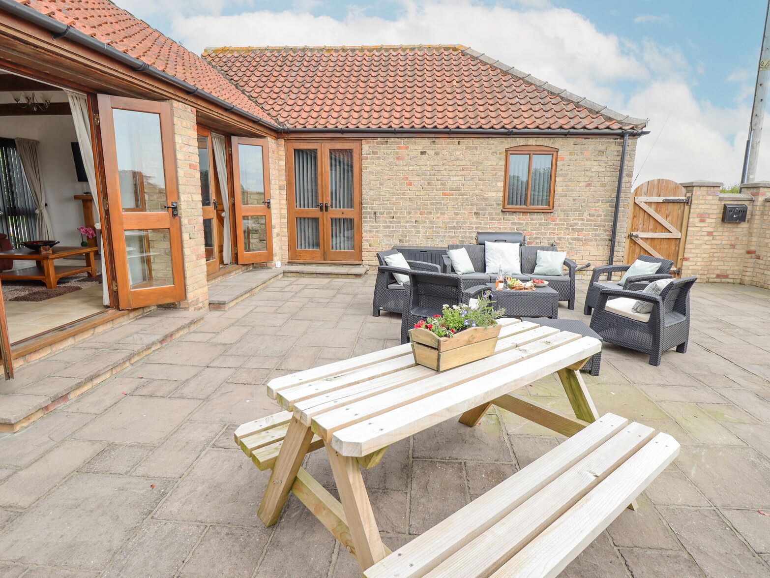 A patio with a wooden table and benches, surrounded by wicker furniture and a brick building with a tiled roof.