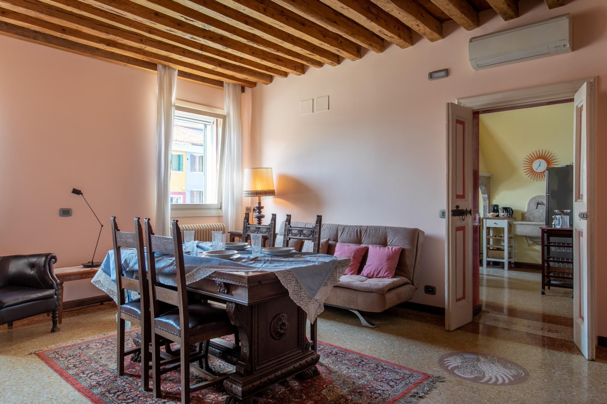 Un comedor con una mesa de madera dispuesta para cuatro personas, un sofá con cojines rosa y una lámpara de estilo tradicional.