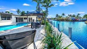 Ein Poolbereich mit Esstisch und Stühlen, einer überdachten Sitzgelegenheit und Blick auf einen Swimmingpool und Palmen.