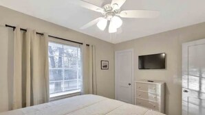 Une chambre à coucher comprenant un lit, un ventilateur de plafond, une télévision, une commode et une fenêtre avec des rideaux.
