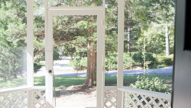 Une véranda couverte avec une balustrade en treillis blanc, un sol en pierre et une vue sur une allée bordée d’arbres.