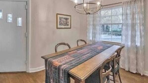 Une salle à manger avec une longue table, des chaises, un lustre, une fenêtre avec des rideaux et une porte.