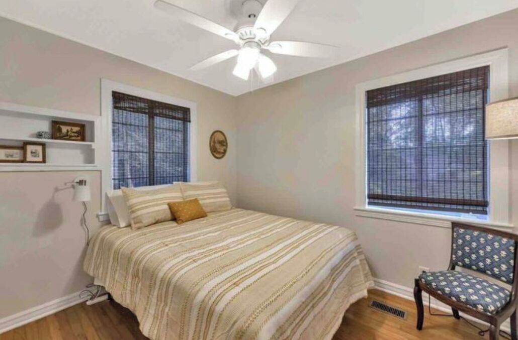 Une chambre à coucher comprenant un lit, une chaise, un ventilateur de plafond et deux fenêtres équipées de stores.